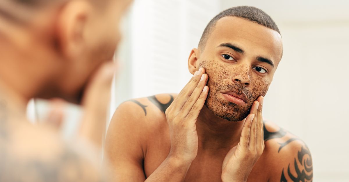 A man rubs an exfoliating scrub on his face while looking in the mirror